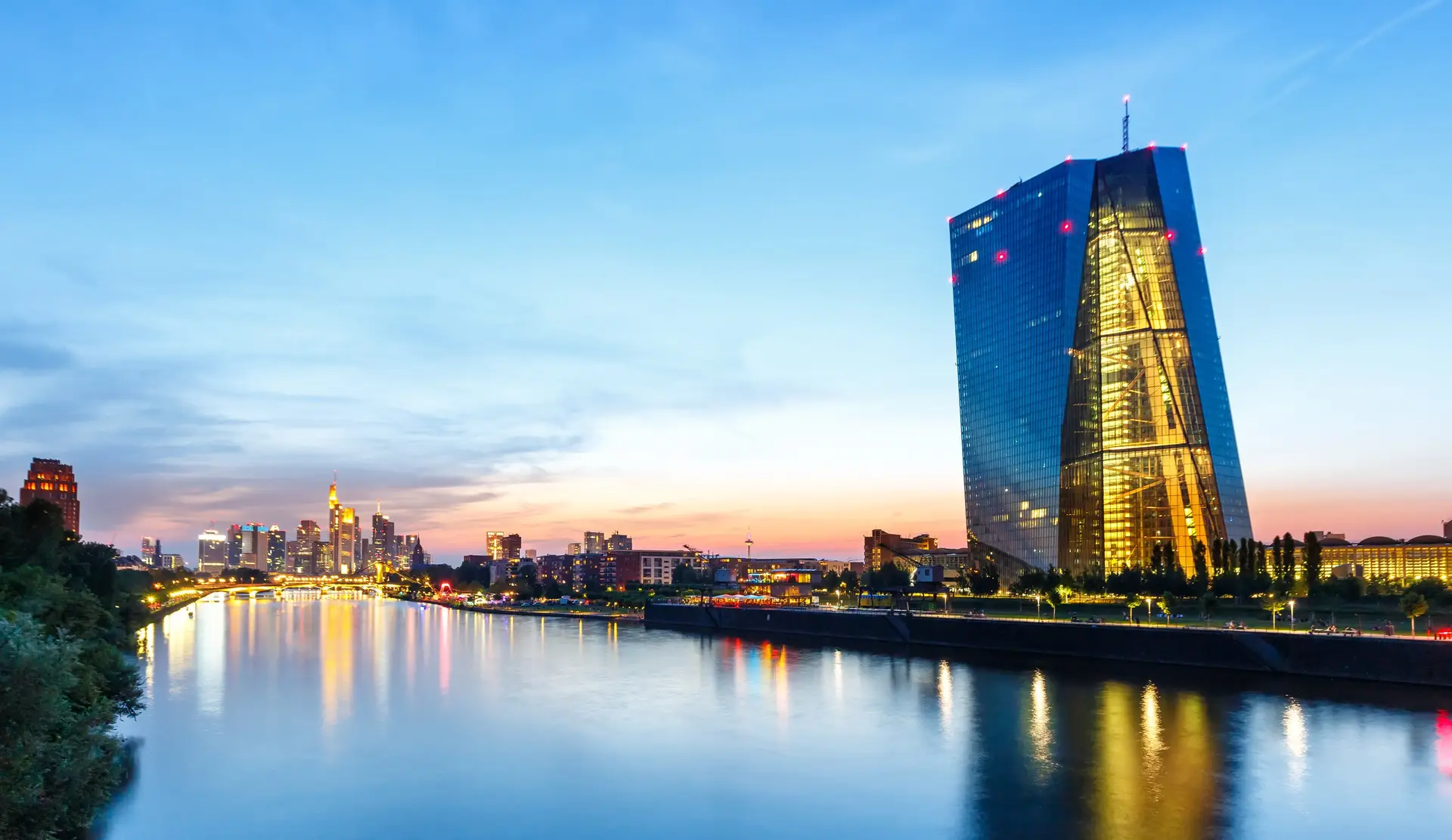 The European Central Bank tower in Frankfurt am Main lit at blue hour, with the city skyline behind it and the river Main in the foreground