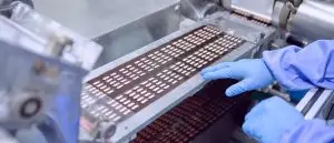 Tablets sealed in blister strips moving along a pharmaceutical packaging line, a gloved hand guiding the strip