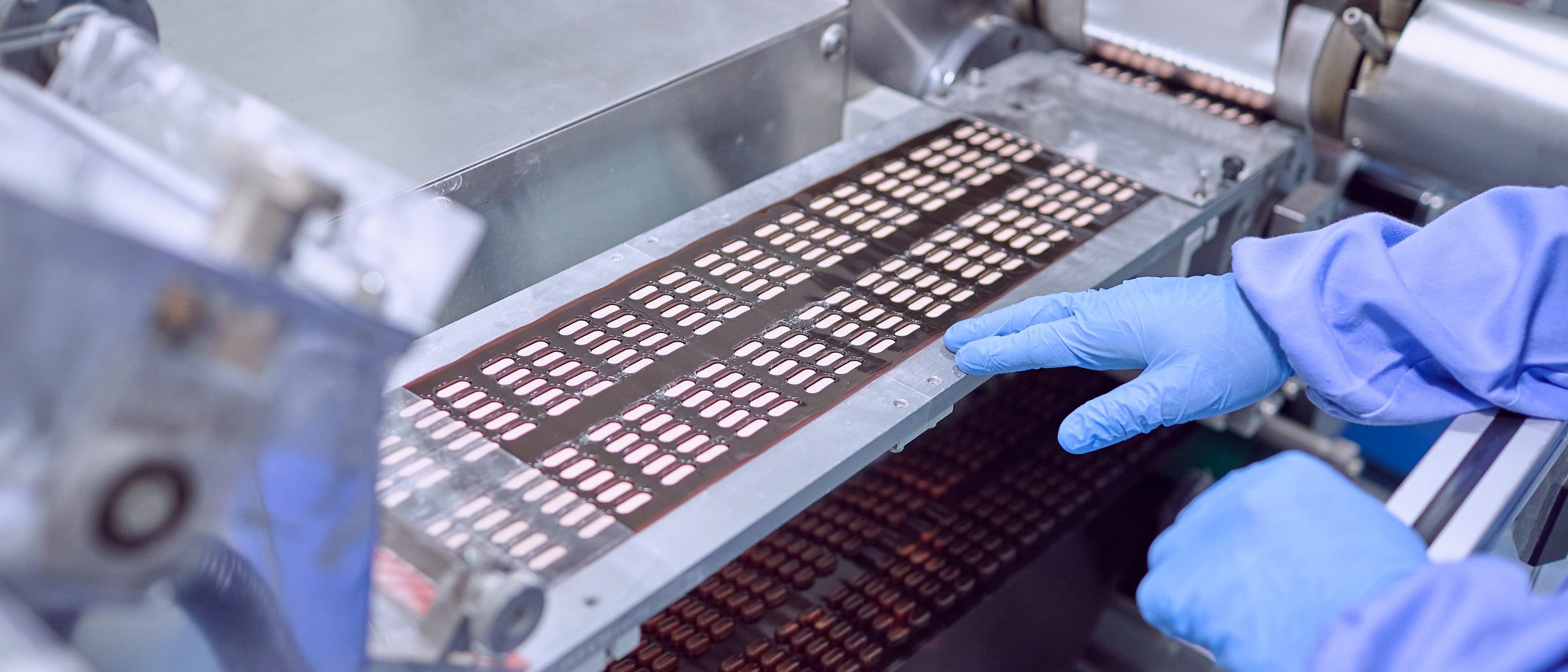 Tablets sealed in blister strips moving along a pharmaceutical packaging line, a gloved hand guiding the strip